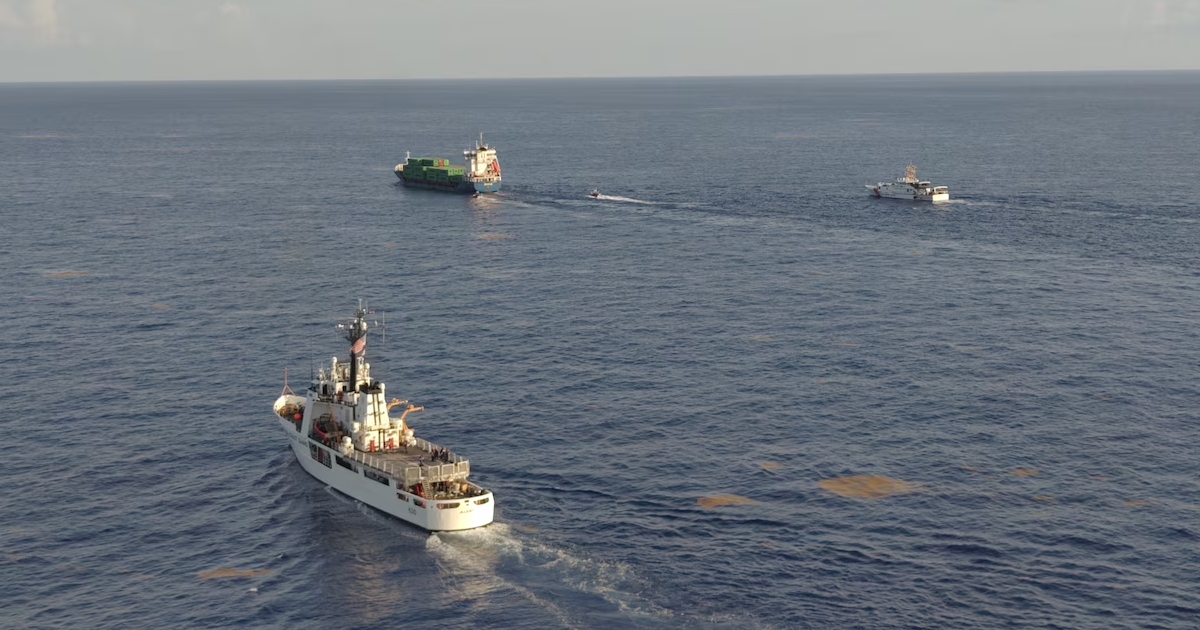 Vista panorámica desde un ángulo elevado que muestra una operación marítima en mar abierto en un día despejado. En primer plano destaca un barco patrullero de la Guardia Costera de Estados Unidos (USCG) navegando en diagonal, dejando una estela blanca en el agua marina. A lo lejos, al centro y hacia la derecha, se observan un buque carguero portacontenedores de casco azul y verde, una lancha de interdicción rápida y otra embarcación oficial guardacostas escoltando al navío.