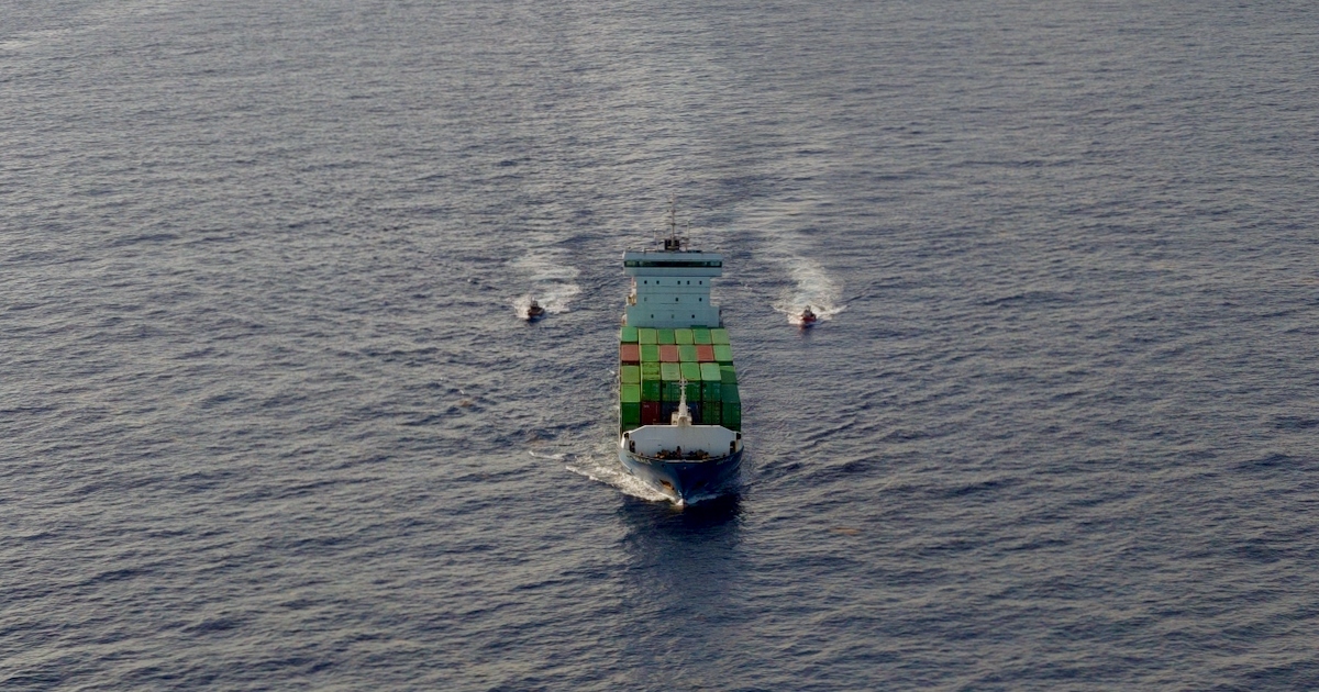 Vista aérea en plano general de un buque portacontenedores navegando en mar abierto. La embarcación lleva contenedores de carga apilados en su cubierta y avanza custodiada por dos embarcaciones de menor tamaño (patrulleras o lanchas de interceptación) que navegan a sus flancos, dejando estelas de agua en la superficie marina.