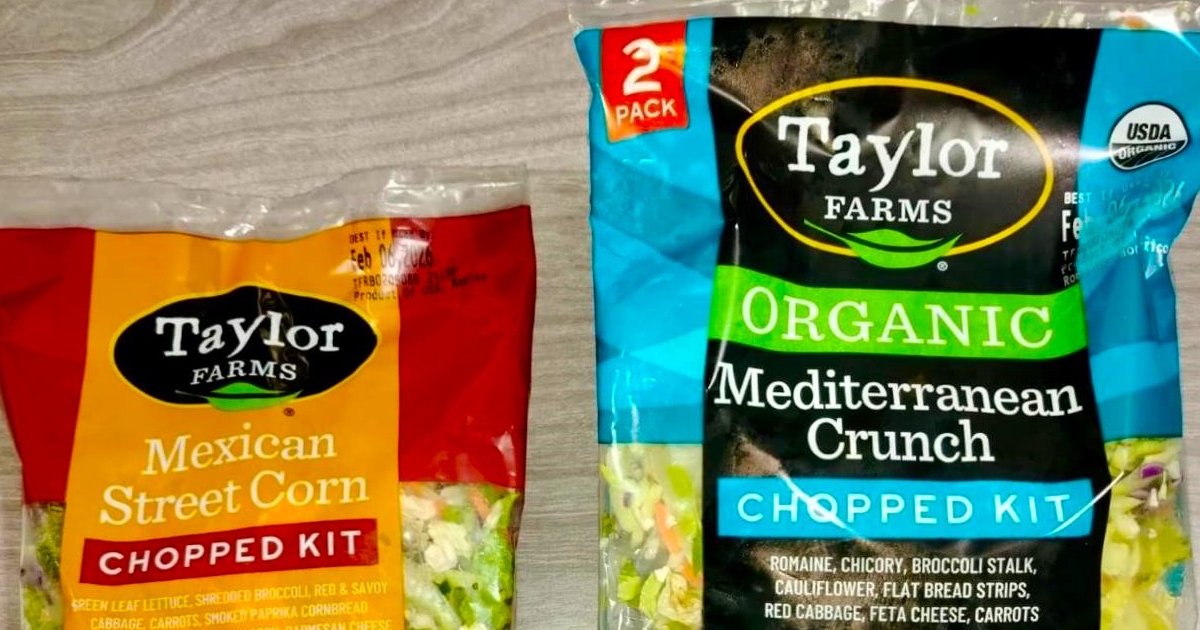 dos empaques plásticos de ensaladas preparadas de la marca Taylor Farms sobre una superficie de madera clara. A la izquierda se observa la bolsa de "Mexican Street Corn Chopped Kit" (de empaque amarillo y rojo), y a la derecha un paquete de dos bolsas ("2 Pack") de "Organic Mediterranean Crunch Chopped Kit" (de empaque azul y verde con sello USDA Organic).